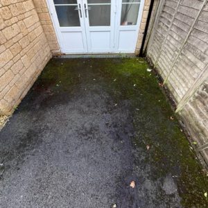 Moss-covered paved area outside white patio doors between a brick wall and fence