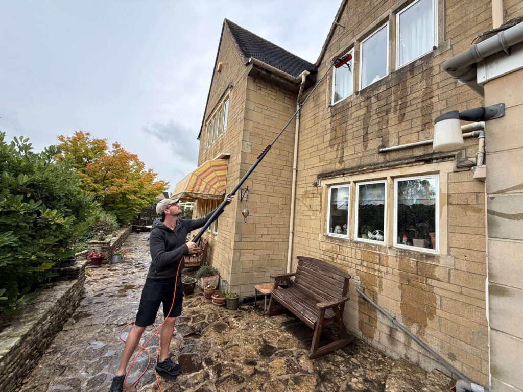 Window cleaner using a pole on first-floor windows of a stone house in Painswick